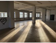 Großer, leerer Industrie- oder Lagerraum mit Betonstützen, hohen Decken und Reihen von Sprossenfenstern, durch die Sonnenlicht lange Schatten auf den Boden wirft. An den Wänden sind Heizkörper und teilweise freihängende Kabel zu sehen.