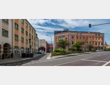 Straßenszene mit moderner Bebauung: Auf der linken Seite ein mehrstöckiges Gebäude mit cremefarbenen und gelben Fassaden sowie grünen Fensterrahmen, rechts ein rosa Gebäude mit der Aufschrift "Potsdamer Straße". Im Vordergrund Verkehrszeichen, eine Bordsteinbegrünung und geparkte Fahrzeuge, im Hintergrund ein wolkiger Himmel.