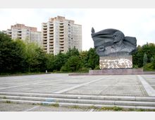 Großer Platz mit grauen Pflastersteinen, im Hintergrund eine monumentale Statue eines Mannes mit erhobener Faust und wehender Fahne, deren Sockel mit Graffiti besprüht ist. Dahinter befinden sich dichte Bäume und hohe Wohngebäude.