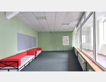 Ein einfacher Raum mit grünen Wänden, einer Reihe roter Sofas entlang der linken Wand, grauen Pinnwänden darüber und einem Poster mit einer Landkarte an der hinteren Wand. Rechts befinden sich große Fenster, durch die Tageslicht einfällt, und der Boden ist mit einem dunkelgrauen Teppich ausgelegt.