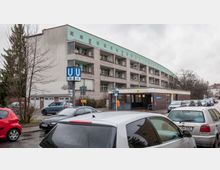 Wohngebäude mit mehreren Balkonen und grünem Flachdach in einer Stadtumgebung, im Vordergrund der Eingang zur U-Bahn-Station "Parchimer Allee" mit blauen U-Bahn-Schildern und geparkten Autos entlang der Straße. Der Himmel ist bewölkt, und kahle Bäume säumen den Rand der Straße.