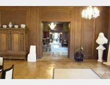 Holzvertäfelter Innenraum eines historischen Gebäudes mit dekorativen Elementen, darunter ein Schrank mit Porzellanvasen, eine weiße Büste auf einem Sockel und ein Kronleuchter an der Decke. Im Hintergrund sind ein weiterer Raum und ein Durchgang mit Teppichen und Möbeln sichtbar.