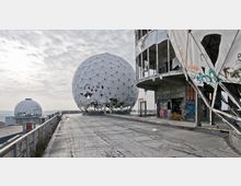 Verlassene ehemalige Abhörstation mit großen, geodätischen Kuppeln aus weißem Stoff, die teilweise beschädigt sind. Im Vordergrund ein grauer Betonweg und ein graffiti-bedecktes, teils zerfallenes Gebäude; im Hintergrund hügelige Landschaft unter bewölktem Himmel.