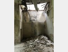 Ein verlassener, heruntergekommener Raum mit beschädigten Wänden und einem eingestürzten Dachbereich, durch den Tageslicht einfällt. Am Boden liegt eine große Menge an Schutt und Trümmern, die auf den Verfall des Gebäudes hinweisen.