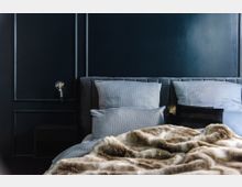 Schlafzimmer mit einem gepolsterten grauen Bettkopfteil, blaugrau gestreiften Kissen, einer Kunstfelldecke und einer dunklen Wand mit schlichter Zierleiste. Links ist eine minimalistische Wandleuchte zu sehen.