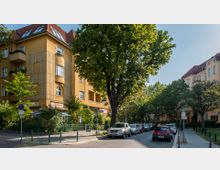 Straßenszene in einem Wohnviertel mit mehrstöckigen Wohnhäusern in gelber Putzfassade und roten Ziegeldächern. Entlang der Straße stehen parkende Autos und zahlreiche Bäume spenden Schatten; im Erdgeschoss eines der Gebäude befindet sich ein Café mit Außensitzbereich.