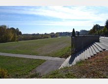 Weite Grünfläche mit Fußwegen, die sich durch die Landschaft ziehen, umgeben von Bäumen im Herbst, im Hintergrund eine Reihe von Gebäuden. Rechts im Vordergrund befinden sich steinerne Treppen mit Geländer, die zu einem höher gelegenen Weg führen.