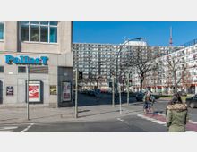 Straßenkreuzung in einer städtischen Umgebung mit einem Gebäude auf der linken Seite, das den Schriftzug „PallasT“ trägt, und einem großen Wohngebäude im Hintergrund. Im Vordergrund ist ein Fußgängerüberweg mit Fußgängern und einem Radfahrer zu sehen, daneben Bäume ohne Laub und Verkehrsampeln.