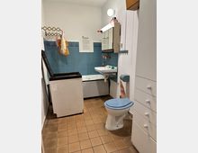 Kleines Badezimmer mit orange-braunen Fliesenboden, blauen Wandfliesen, Waschbecken und Spiegelschrank. Eine Toilette mit blauem Sitz, eine Badewanne und ein altes Gerät, vermutlich eine Waschmaschine, sind ebenfalls zu sehen.