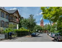 Wohnstraße mit parkenden Autos, gesäumt von grünen Bäumen und einem Zaun, links ein zweigeschossiges Gebäude im traditionellen Stil mit Holzdetails. Im Hintergrund ragt ein roter Kirchturm mit grünem Spitzdach über die Szene hinaus.