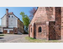 Backsteinbau einer historischen Kirche mit Spitzbogenfenstern und dekorativen Ziegelmustern, daneben ein weißes Gebäude mit roten Ziegeldetails. Der gepflasterte Platz ist von Bäumen gesäumt, bei sonnigem Wetter.