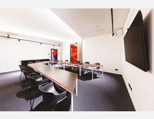 Moderner Konferenzraum mit weißen Wänden, einem U-förmigen Tisch mit schwarzen Stühlen und einem großen Bildschirm an der rechten Wand. Im Hintergrund befindet sich eine Glastür mit roten Akzenten sowie eine Glasfläche mit roter Beleuchtung.