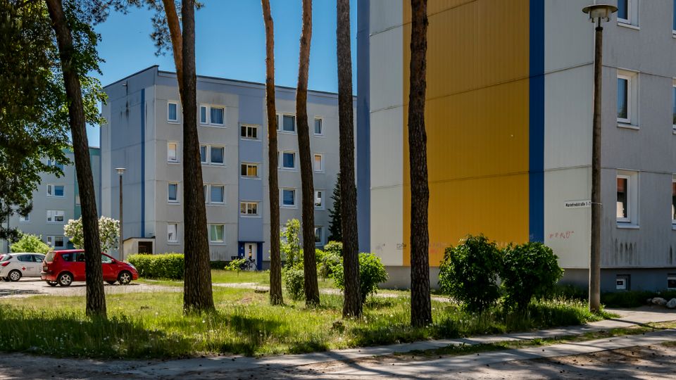 Wohnanlage mit mehrstöckigen, flachgedeckten Wohnblocks in Grau und Gelb, umgeben von einer grünen Rasenfläche mit hohen Bäumen. Im Vordergrund verläuft ein Gehweg; daneben stehen Straßenlampen, geparkte Autos und Fahrräder am Gebäudeeingang.