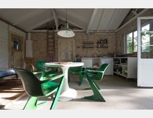 Innenraum eines rustikalen Wohnbereiches mit offener Küche, sichtbaren Ziegelwänden und weißem Dachstuhl. Im Vordergrund steht ein weißer runder Tisch mit grünen Designerstühlen, dahinter eine einfache Küchenzeile mit offenen Regalen und einem Fenster, das Tageslicht hereinlässt.