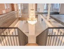 Innenraum eines sanierten Industriegebäudes mit einer hölzernen Treppe und Metallgeländern. Im Zentrum hängt ein großer, kristallener Kronleuchter; Wände aus Backstein und große Fenster prägen den Raum, ergänzt durch weiße Säulen und Metallelemente.