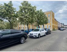 Straßenszene mit parkenden Autos am Straßenrand vor einem gelb gestrichenen Gebäude mit mehreren Stockwerken und weißen Fensterrahmen. Im Hintergrund stehen blühende Bäume und weitere mehrfarbige Gebäude entlang der Straße.