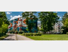 Mehrstöckige Wohngebäude in einer Wohnsiedlung, umgeben von Bäumen und Grünflächen. Eines der Gebäude hat eine farbenfrohe, abstrakte Fassadengestaltung in Orange, Blau und Weiß.
