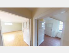 Wohnungsflur mit zwei sich öffnenden weißen Türen, die in verschiedene Räume führen. Links befindet sich ein Raum mit hellem Holzboden und kleinem Fenster, rechts ein Raum mit braunen Fliesenboden und einem größeren Fenster.