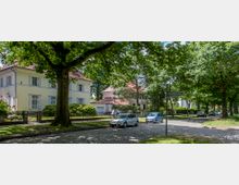 Straßenszene in einem grünen Wohngebiet mit gepflasterter Straße, großen Bäumen und Einfamilienhäusern mit hellem Putz und roten Ziegeldächern; parkende Autos sind am Straßenrand zu sehen. Ein Straßenschild mit der Aufschrift "Blumenthalstraße" steht im Vordergrund.