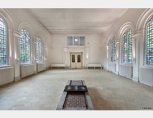 Heller Innenraum mit hohen, farbigen Glasfenstern und Rundbögen an den Wänden, einem hellen Fliesenboden und einem zentralen bodennahen Steinbrunnen. Im Hintergrund sind ein paar weiße Sitzbänke und eine Doppeltür mit Glasscheiben zu sehen.