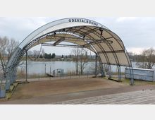 Freiluftbühne mit gewölbtem Dach und Metallträgern, beschriftet mit "Odertalbühne", im Vordergrund eine Holzplattform mit angrenzender Pflasterfläche. Im Hintergrund ist ein Fluss mit kahlen Bäumen und einer ländlichen Umgebung zu sehen.
