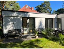 Hinterhof eines modernen Einfamilienhauses mit weißen Putzwänden und großen Glas-Schiebetüren, die zu einer Holzterrasse führen. Auf der Terrasse stehen Holzbänke, ein Tisch mit Stühlen und Pflanzentöpfe; im Hintergrund sind Bäume und ein weiteres Gebäude mit rotem Ziegeldach sichtbar.
