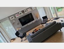 Modernes Wohnzimmer mit hellen Holzböden, einer grauen Couch und einem großen Wandfernseher. An der Wand befinden sich Regale mit Bilderrahmen, darunter ein graues Sideboard; große Fenster und weiße Vorhänge lassen viel Tageslicht herein.