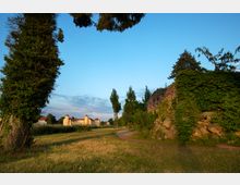 Weitläufige Parklandschaft mit einem grasbewachsenen Weg, der an einer mit Efeu bewachsenen Steinmauer entlangführt; im Hintergrund ein helles, historisches Gebäude mit Türmen und roten Dächern, umgeben von Bäumen und Wiesen unter klarem, blauem Himmel.