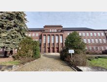 Backsteinbau mit symmetrischer Fassade und hohen Fenstern, flankiert von Bäumen und Büschen. Ein gepflasterter Weg führt zu einer Treppe und mehreren Türen, mit einem Schild im Vordergrund, das „Privatgrundstück – Betreten verboten“ anzeigt.