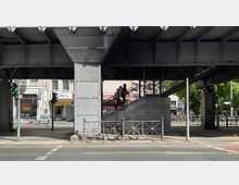 Straßenszene unter einer erhöhten Bahntrasse aus Stahl mit sichtbaren Trägern und Pfeilern. Im Vordergrund ein Gehweg mit einem Geländer und geparktem Fahrrad, während eine Person eine Treppe hinaufgeht; im Hintergrund befinden sich Gebäude mit Geschäften und Graffiti.