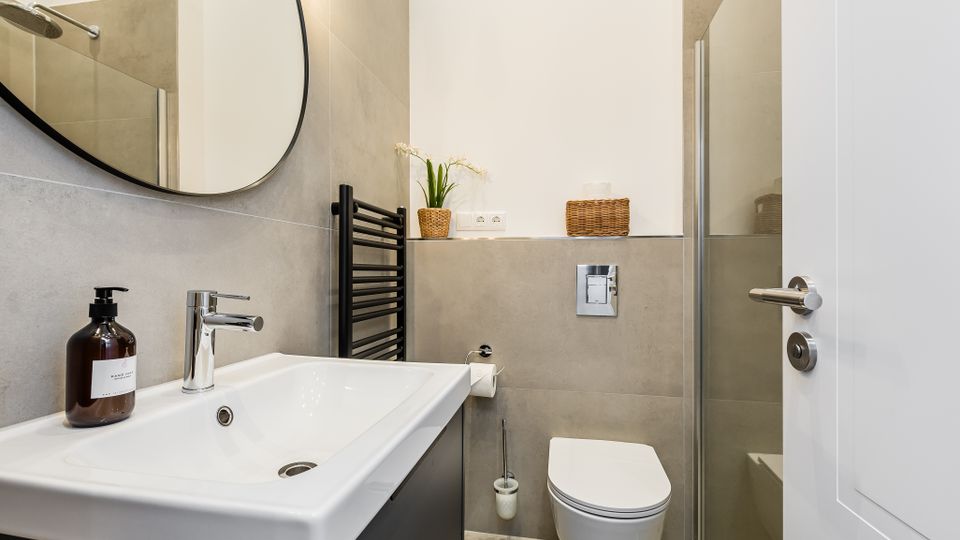 Modernes Badezimmer mit grauen Fliesen, weißem Waschtisch mit rundem Spiegel und wandhängendem WC. Rechts ist eine Glasdusche zu sehen, dazu ein schwarzer Handtuchheizkörper und kleine Körbe sowie eine Pflanze als Ablageelemente.