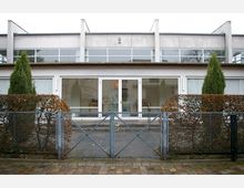 Moderne Wohnhausfassade mit großen Glasfronten im oberen und unteren Bereich, flankiert von symmetrisch angeordneten Hecken und zwei Zierbäumen. Ein Metallzaun mit einem Tor begrenzt den Vorderbereich des Hauses.