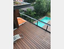 Terrasse aus Holz mit einem Liegestuhl und einer Matratze mit orangefarbener Decke, umgeben von Bäumen. Im Hintergrund befindet sich ein rechteckiger Swimmingpool mit Leiter sowie ein Garten mit dichter Vegetation.