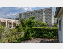 Mehrstöckiges Wohngebäude mit Balkonen im Hintergrund, umgeben von einer begrünten Gartenanlage mit Büschen und Bäumen. Im Vordergrund ein Maschendrahtzaun, neben einem flachen Gebäude mit großen Fenstern.