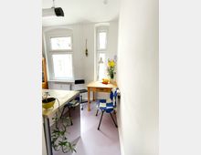 Kleiner, heller Raum mit zwei Fenstern, einem Holztisch und einem Stuhl mit blau-weißem Muster. Auf dem Tisch stehen eine Tischlampe, eine Vase mit Blumen und einige Gegenstände, während im Vordergrund eine Pflanze auf einer Arbeitsfläche zu sehen ist.