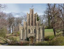 Backsteingebäude im neogotischen Stil mit Spitzbogenfenstern und dekorativen Elementen, umgeben von kahlen Bäumen und einer Wiese in ländlicher Umgebung. Ein geschwungener Weg führt zu einem Torbogen, der von Grünpflanzen umrahmt ist.