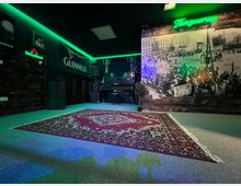 Innenraum einer Bar oder eines Veranstaltungsraums mit dunklen Holzpaneelen, grünem LED-Deckenlicht und Leuchtschildern. Im Hintergrund stehen ein Klavier mit Lautsprechern, an der Wand hängt ein großes Schwarzweiß-Fotomural, und auf dem Boden liegt ein großer gemusterter Teppich.