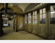 Langer, schmaler Flur in einem historischen Gebäude mit gewölbter Decke, alten Holztüren mit Glaseinsätzen und sichtbaren Gebrauchsspuren. Rechts führen Treppen hinter den Glastüren nach oben, während im Hintergrund eine weitere Holztür den Flur abschließt.