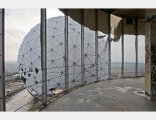 Ansicht einer großen, weißen, geodätischen Kuppel in ruinöser Umgebung, aufgenommen von einem offenen Betonbau mit Graffiti. Im Hintergrund ist eine bewaldete Landschaft unter einem bewölkten Himmel zu sehen.