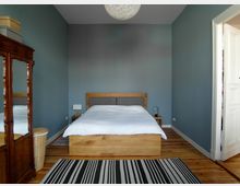 Schlafzimmer mit blauen Wänden, einem Holzbett mit weißer Bettwäsche und zwei Nachttischen mit Leselampen. Links steht ein antiker Kleiderschrank mit Spiegel, der Boden ist aus Holz, und ein gestreifter Teppich liegt vor dem Bett.