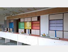 Innenraum eines verlassenen Klassen- oder Laborraums mit langen Arbeitstischen und mehreren Wasserhähnen; an der Rückwand stehen Einbauschränke und Regale mit farbigen Glasfronten. Die Decke zeigt abblätternde Farbe und beschädigten Putz.