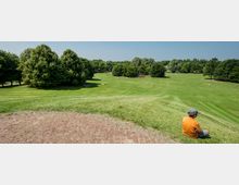 Weitläufige Parklandschaft mit einer großen, offenen Wiese, gesäumt von dicht bewachsenen Bäumen; im Vordergrund sitzt ein Kind mit orangem Shirt auf dem grasbewachsenen Hang, während im Hintergrund vereinzelt Menschen auf der Wiese entspannen.