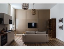 Modern eingerichtetes Wohnzimmer mit offener Küche. Ein beiges Sofa steht vor einer Akzentwand mit Fernseher, links befindet sich eine Küchenecke mit Einbaugeräten, rechts ein hölzernes Regal, und an der Wand hängen gerahmte Bilder.