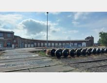 Historisches Eisenbahndepot mit einem langgezogenen, halbkreisförmigen Backsteingebäude mit großen Toren und Fenstern; im Vordergrund Schienen und aufgereihte Eisenbahnräder auf Holzpaletten. Der Himmel ist leicht bewölkt.