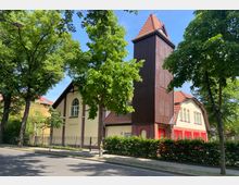 Kirchengebäude mit cremefarbenen Wänden, burgunderroten Akzenten und einem hohen, hölzernen Kirchturm mit rotem Ziegeldach. Im Vordergrund stehen Bäume und ein Zaun, die das Gebäude teilweise umgeben; eine Straße führt daran vorbei.