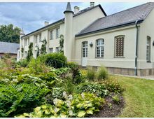 Hellfarbenes, zweigeschossiges Landhaus mit Satteldach, mehreren Kaminen, weißen Sprossenfenstern und einem runden Eckturm; an der Fassade wachsen Kletterpflanzen. Davor liegt ein gepflegtes Gartenbeet mit dichtem Grün und Stauden neben einem Rasenstreifen.