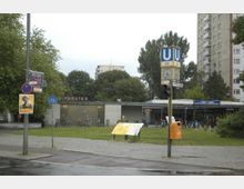 Außenansicht eines U-Bahnhofs mit dem Schild „U9“ und der Bezeichnung „Bartholdyplatz“. Im Hintergrund ein modernes Gebäude mit der Aufschrift „Theater“, umgeben von Bäumen, Fahrrädern und einem Gehweg. Rechts sind Hochhäuser sichtbar.