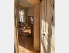 Blick durch eine geöffnete, verzierte Holztür in einen alten Salon mit hohen Fenstern, Parkettboden und einem Kronleuchter. Im Raum stehen ein kleiner runder Tisch, ein gepolsterter Sessel und ein Spiegel; die Wände wirken abgenutzt und teilweise abgeblättert.