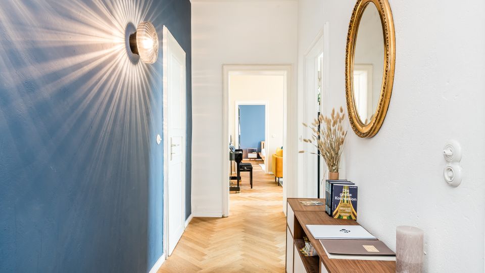 Heller Flur in einer Wohnung mit blau gestrichener Akzentwand, Wandlampe mit Strahlenlicht, weiß gestrichenen Türen und Fischgrätparkett. An der rechten Wand hängen ein runder Spiegel mit Goldrahmen und darunter steht ein schmaler Konsolentisch mit Deko; im Hintergrund führt der Flur in einen Wohnraum.
