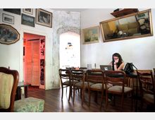 Ein gemütlicher Innenraum mit Holzböden, einer langen Holztafel mit Stühlen und an der Wand hängenden Bildern und Spiegeln. Im Hintergrund befinden sich eine Nische mit heller Steinwand und eine Tür mit roten Lichtakzenten, während eine Frau an einem Laptop arbeitet.